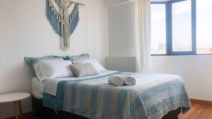 Ein ordentlich bezogenes Bett mit einer blau-weißen gestreiften Tagesdecke, ein Wandbehang aus Makramee und Blick auf die Stadt durch ein großes Fenster mit weißen Vorhängen.
