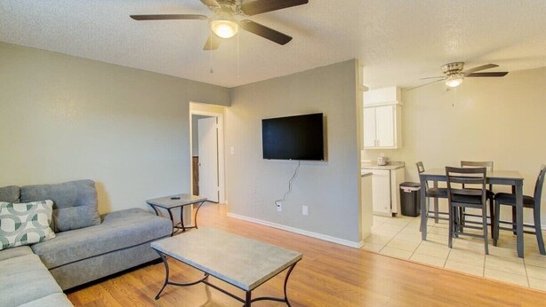 A living room with a sofa, coffee table, and a TV mounted on the wall. There is a dining area with a table and chairs.