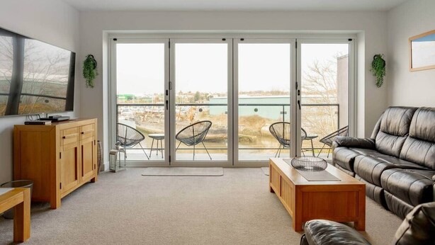 A modern living room with a large sliding glass door leading to a balcony, a leather sofa, a wooden coffee table, and a TV on a wooden cabinet.