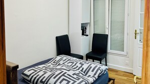 Une chambre à coucher comprenant un lit, deux chaises, une armoire, un luminaire au plafond et une fenêtre avec des stores.