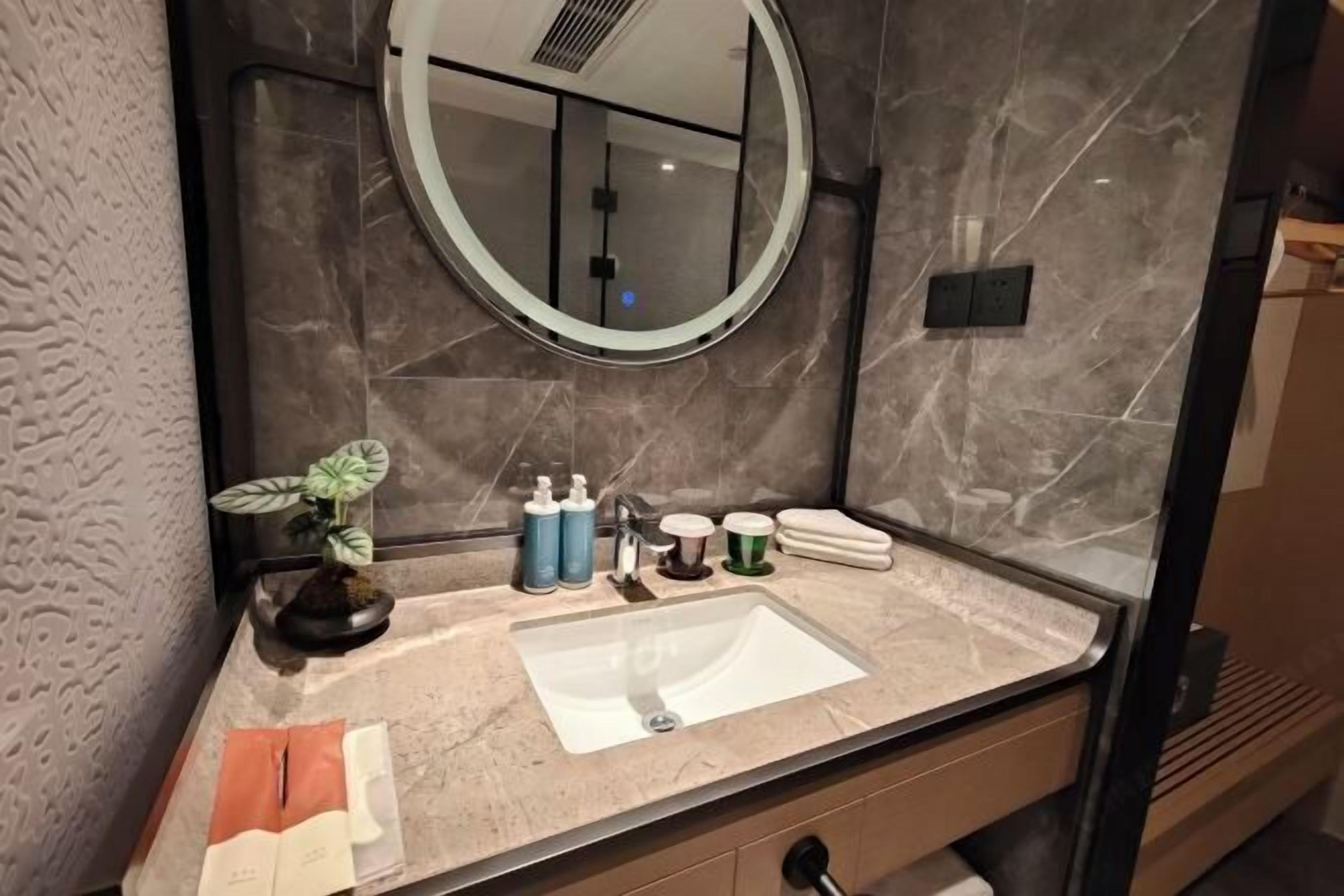 A bathroom with a marble sink, a round mirror, toiletries, and a potted plant.