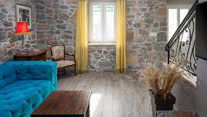 Une pièce avec un mur en pierre, un canapé bleu, une table en bois, une chaise et une fenêtre avec des rideaux jaunes.