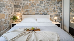 Une chambre avec un mur en pierre, un lit blanc, deux tables de chevet avec des lampes et un plateau sur le lit, sur lequel se trouvent une poire et une tasse.
