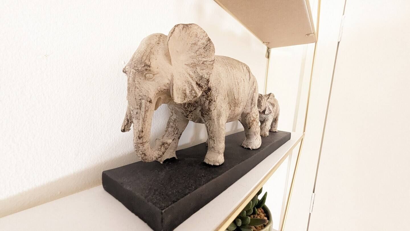 A sculpture of an elephant and a calf on a black shelf against a white wall.