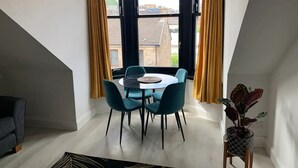 Een eetruimte met een ronde tafel en vier stoelen, een potplant en uitzicht op gebouwen door grote ramen met gordijnen.