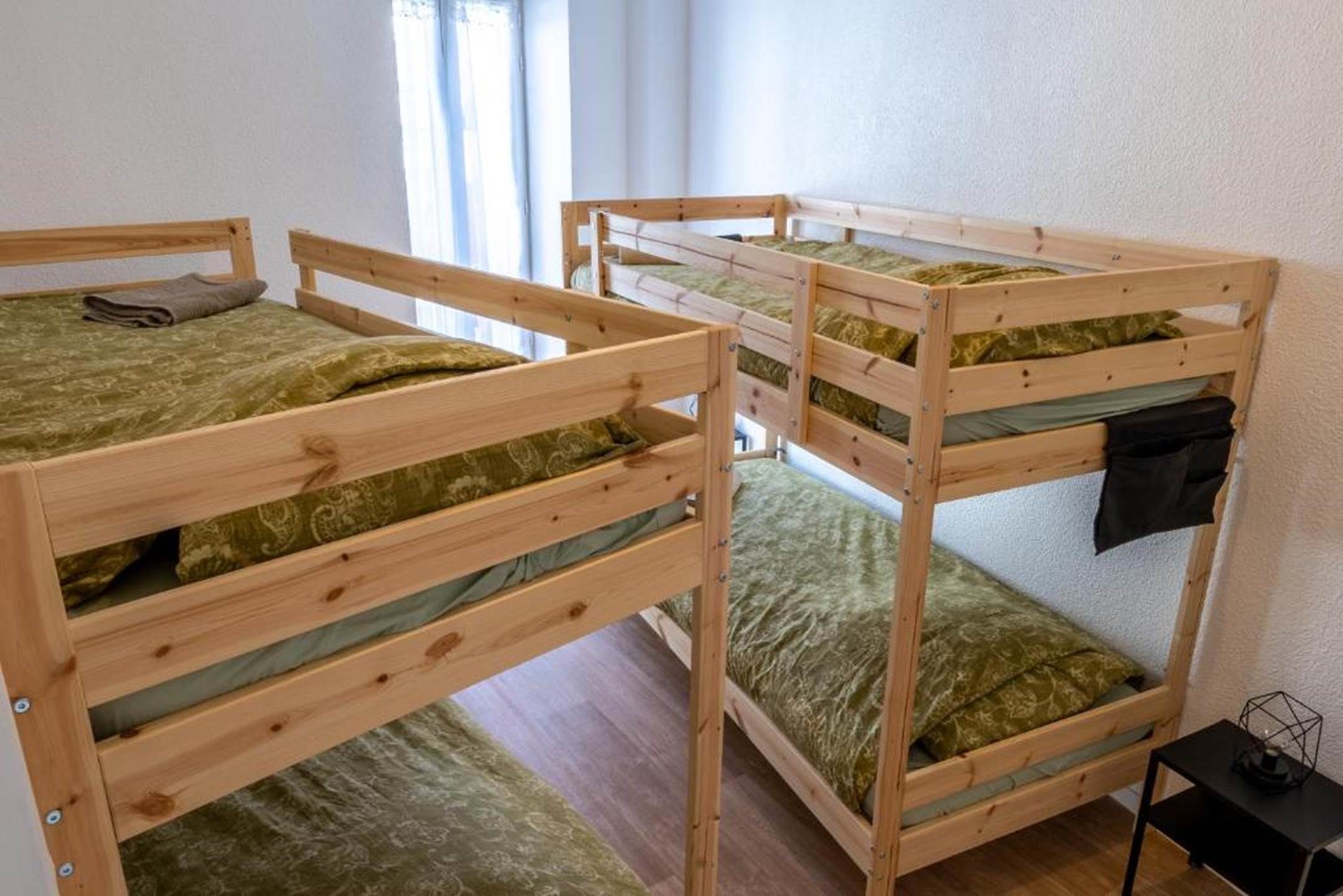 A room with three bunk beds, each with a green patterned mattress, a wooden frame, and a small table with a lamp beside it.