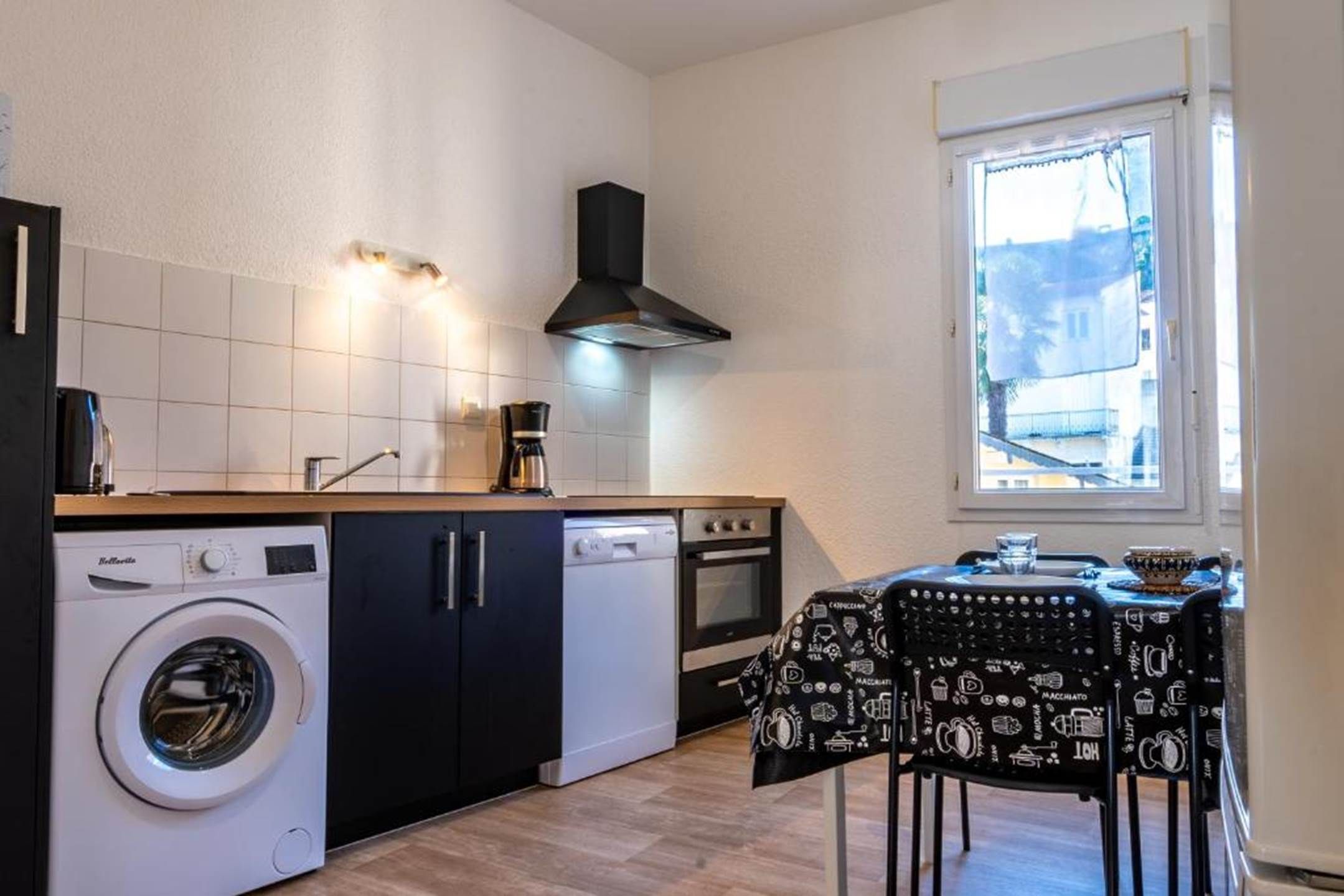 A compact kitchen with a washing machine, a coffee maker, and a dining table with a black tablecloth featuring coffee and tea illustrations.