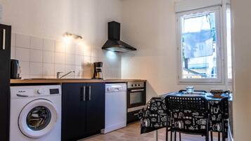 A compact kitchen with a washing machine, a coffee maker, and a dining table with a black tablecloth featuring coffee and tea illustrations.