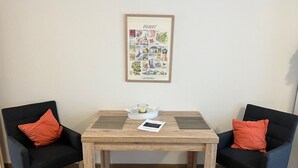 Une pièce avec une table en bois, deux chaises noires dotées de coussins orange, un tableau encadré accroché au mur et un petit bol blanc posé sur la table.