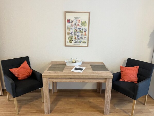 Una habitación con una mesa de madera, dos sillas negras con cojines naranja, un cuadro en la pared y un cuenco blanco pequeño sobre la mesa.