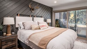 Une chambre moderne avec un grand lit, deux lampes de chevet, une tête de lit en bois ornée de bois de cerf, et une vue sur les arbres à travers une grande fenêtre.