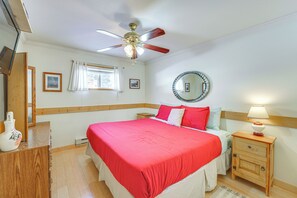 Une chambre à coucher comprenant un lit, une table de chevet en bois, une lampe de chevet, un ventilateur de plafond, une fenêtre avec des rideaux et un miroir sur le mur.