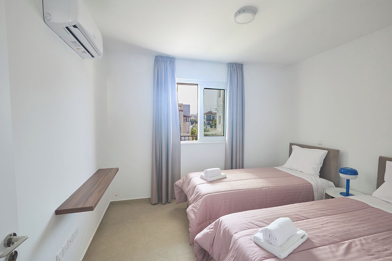 Hotellihuone, jossa on kaksi sänkyä, ikkuna, jossa on verhot, yöpöytä lampun kanssa ja ilmastointilaite.