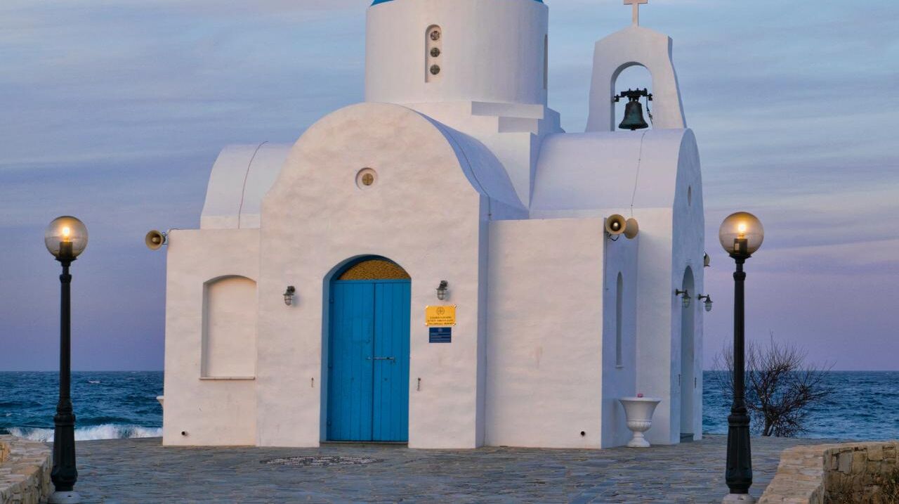 Una iglesia blanca con puerta azul y un campanario, cerca del mar.