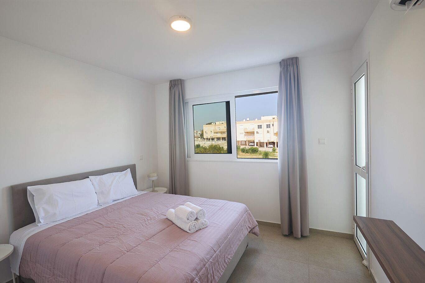 A hotel room with a bed, a nightstand, a window with curtains, and a view of buildings outside.