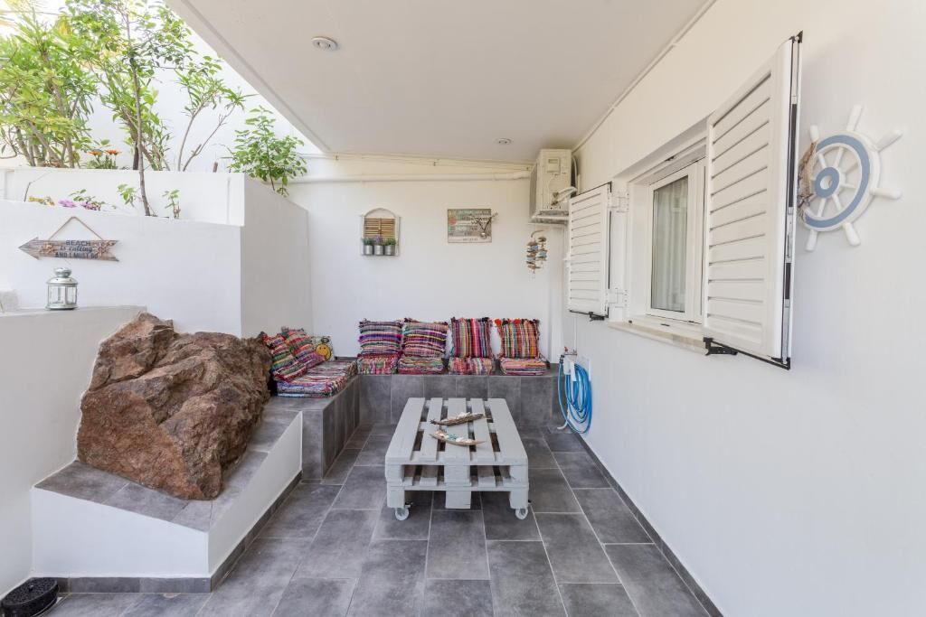 Een klein, afgesloten terras met een witte muur, een grote rots, kleurrijke kussens, een houten pallet en een decoratie van een scheepsschroef.