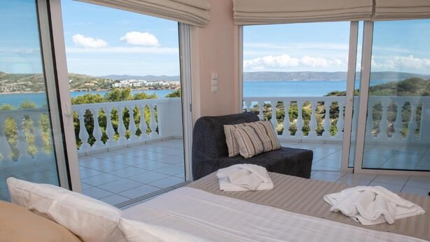 A bedroom with a bed, a chair, and a view of the sea and hills. - Sounio Golden View Villas (Lavreotiki)