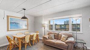 Ein Wohnzimmer mit einem beigen Ledersofa, einem Esstisch mit Korbstühlen, einem gerahmten Gemälde und Blick auf Gebäude durch ein großes Fenster.