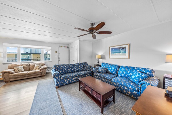 A living room with a beige sofa, a blue patterned sofa, a wooden coffee table, and a ceiling fan. - Sea Horse 306 by Seaside Vacations & Sales (Ocean City)