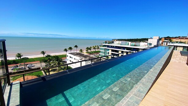 A long, clear swimming pool with a view of the beach and ocean. - Flat FM20 Cabo Branco L3 - By Almare (João Pessoa)