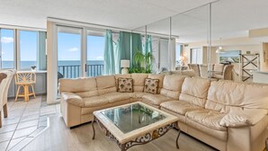 Un amplio salón con un gran sofá seccional color beige, una mesa de centro de vidrio y vistas al océano a través de grandes ventanales.