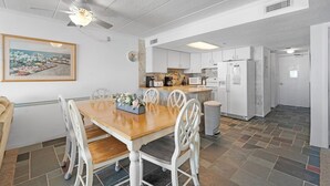 Une cuisine moderne avec des meubles blancs, un coin repas doté d’une table en bois et de chaises blanches, et un ventilateur de plafond.