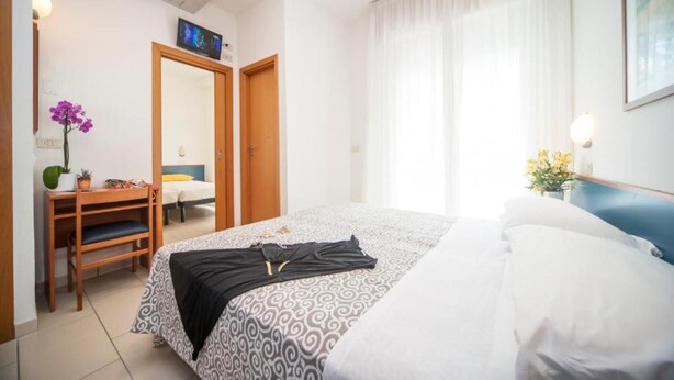 A hotel room with a bed, a desk with a chair, a mirror, and a television. - HOTEL TAORMINA (Riccione)