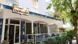 A hotel named 'Hotel Taormina' with a covered entrance, outdoor seating, and a tree in the foreground. - HOTEL TAORMINA (Riccione)
