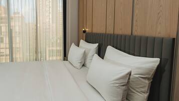 Moderni hotellihuone, jossa on suuri sänky, puinen pääty ja näkymä kaupunkiin läpikuultavien verhojen läpi.