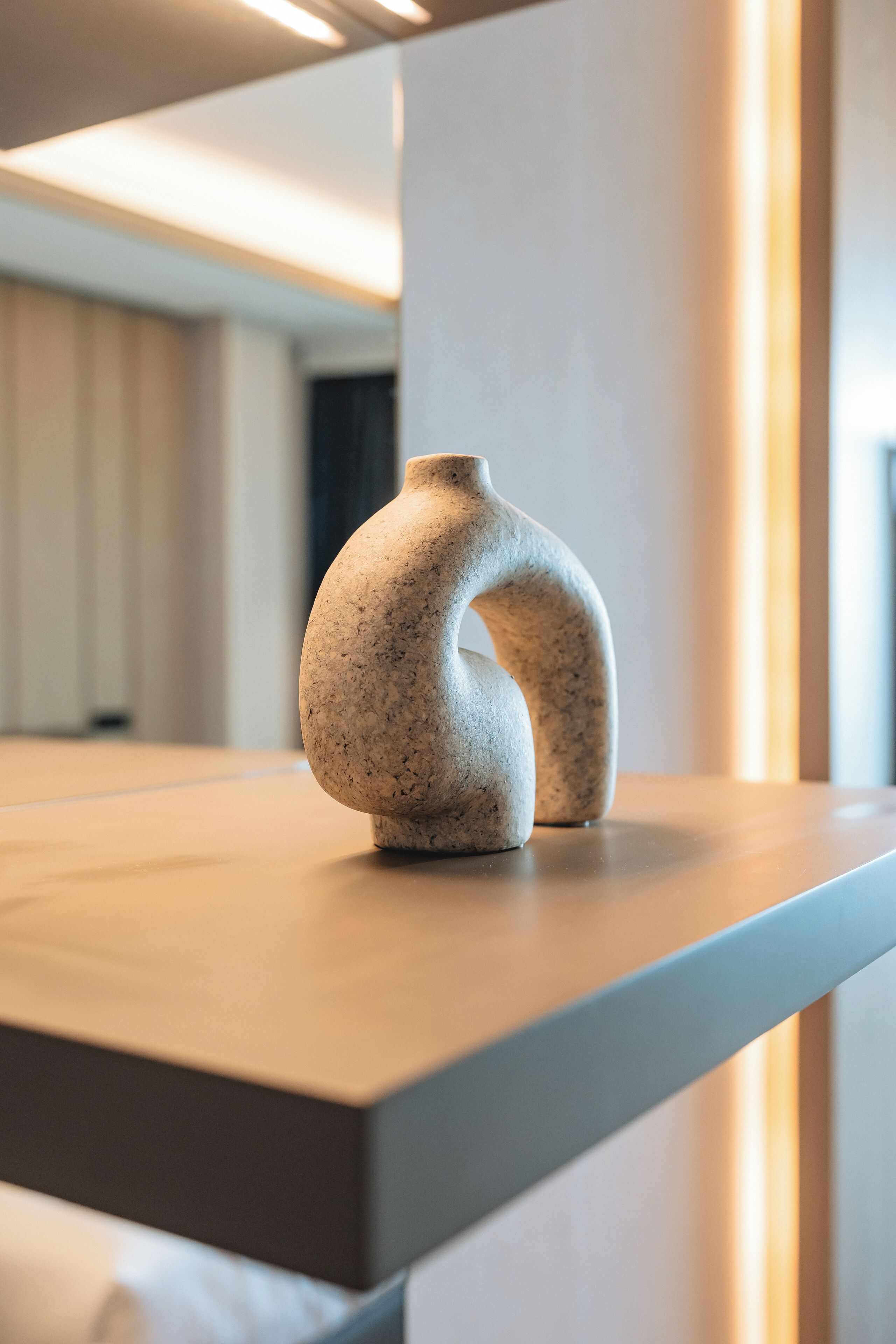 A modern, minimalist vase on a wooden surface in a well-lit room with a neutral color palette.