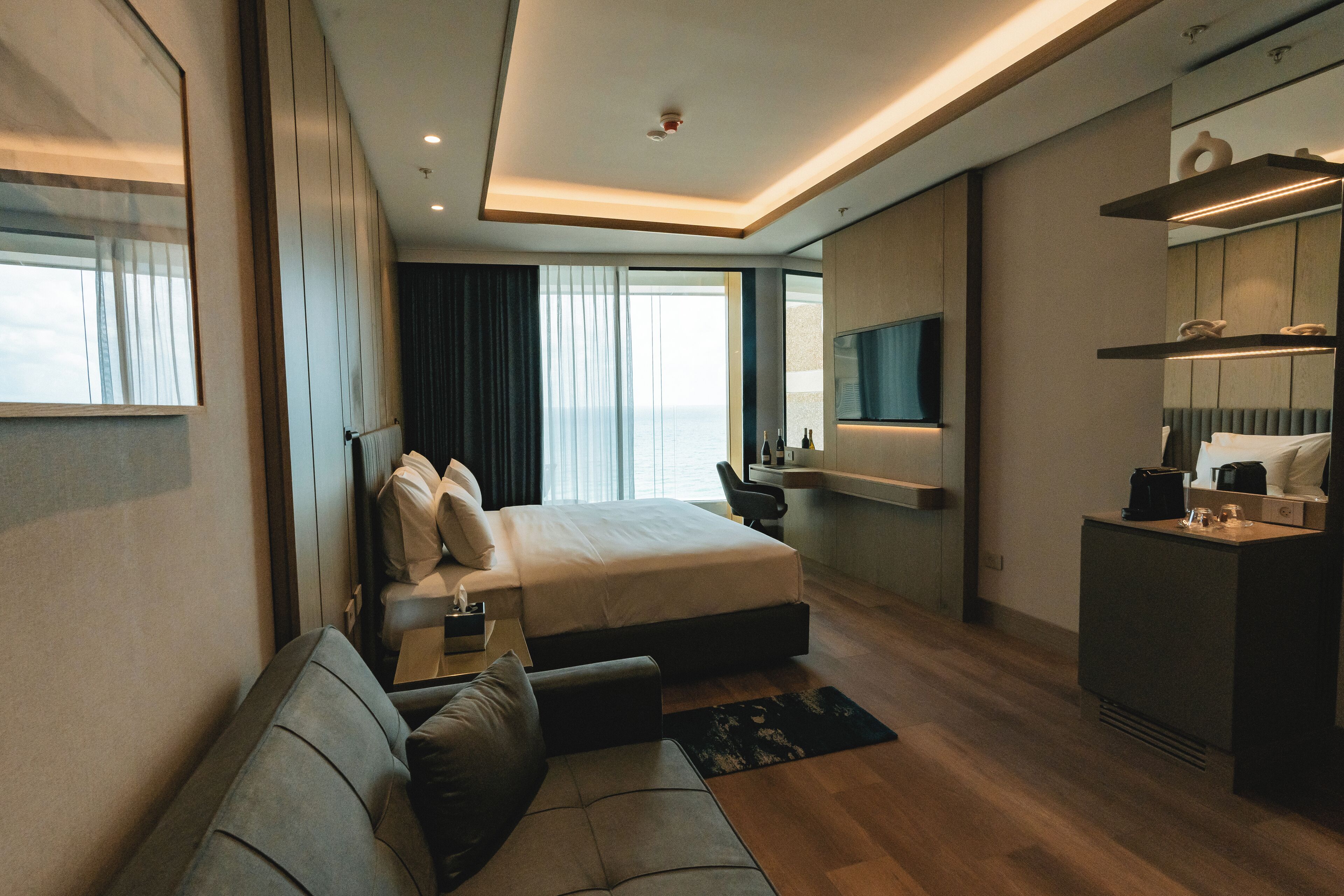 Una habitación de hotel moderna con una cama grande, un sofá, un escritorio con una silla, un televisor montado en la pared y una pequeña kitchenette.