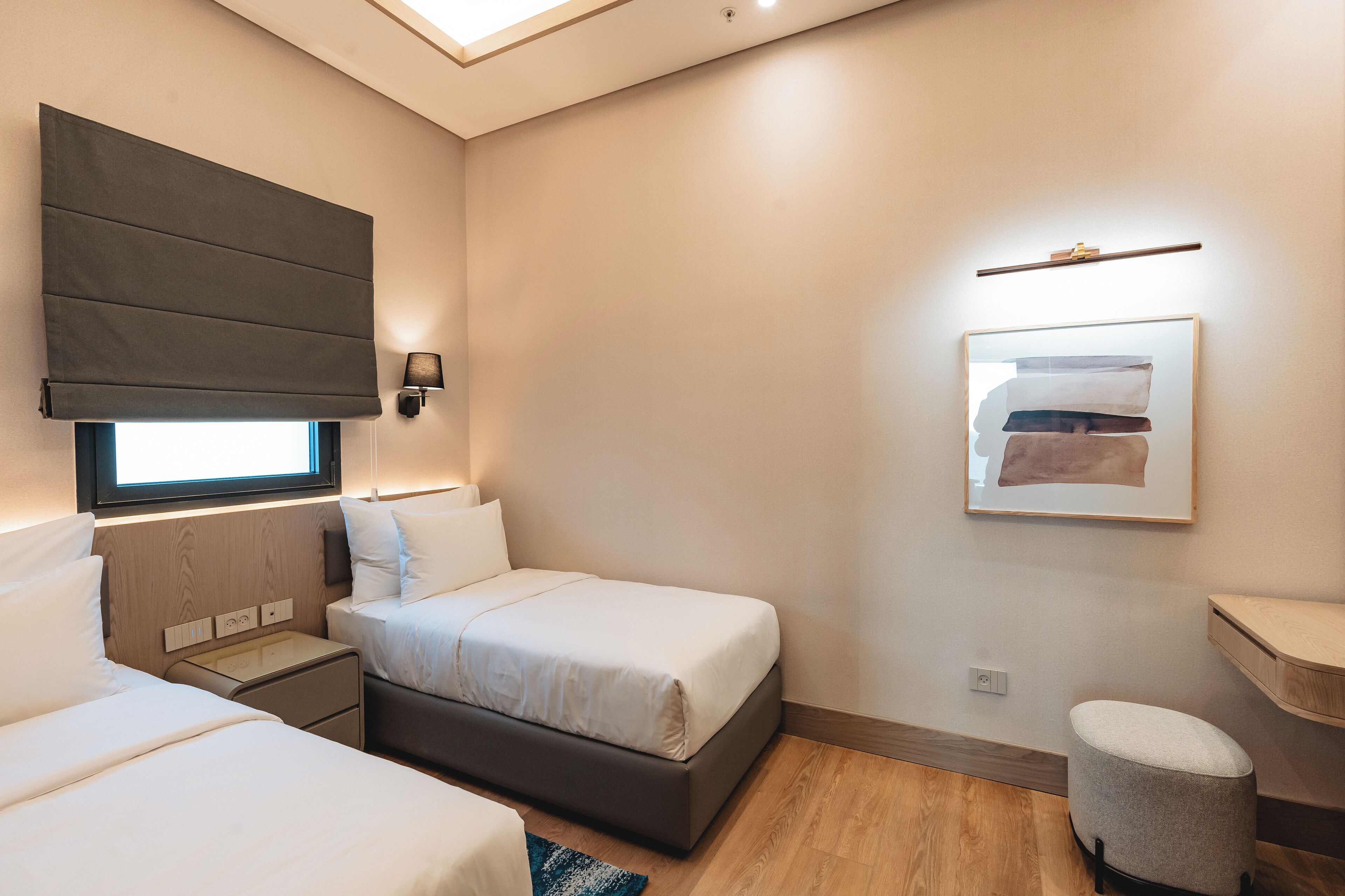 Habitació d'hotel moderna amb dos llits, una taula de nit, un petit tamboret, una finestra amb vista i una obra d'art penjada a la paret.