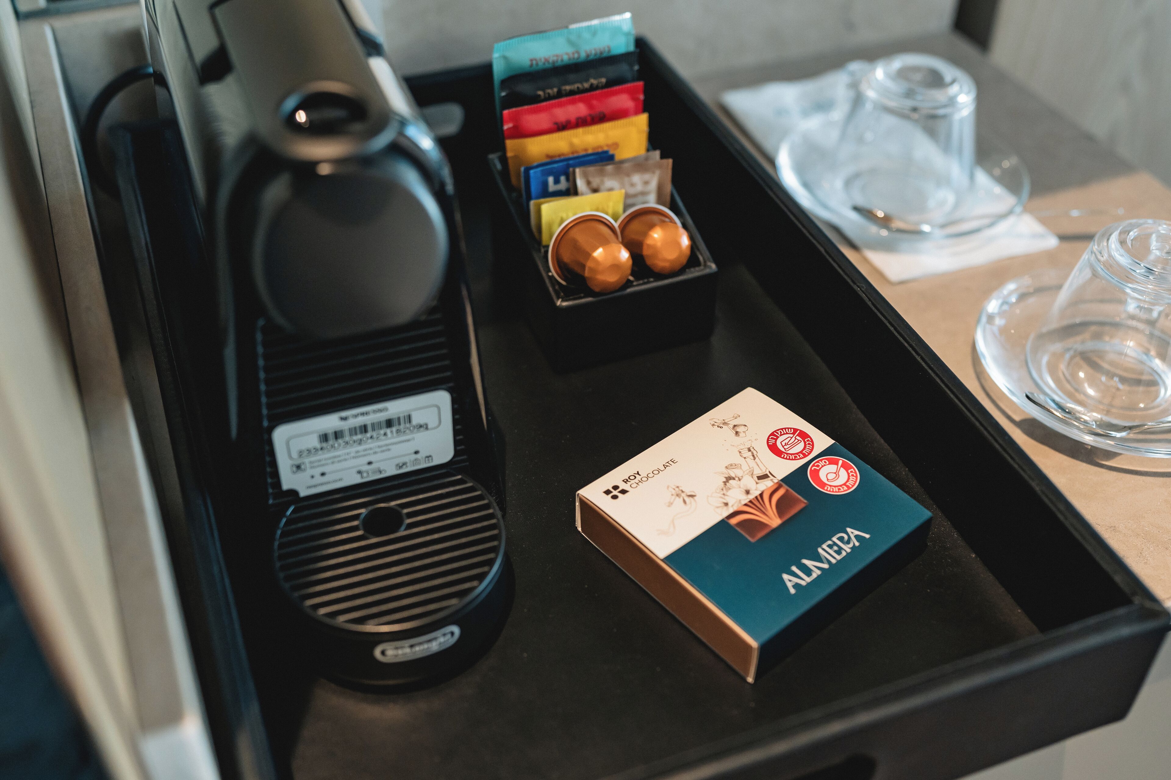 Una bandeja con una caja de bombones Almena, una cafetera y un juego de cápsulas de café.
