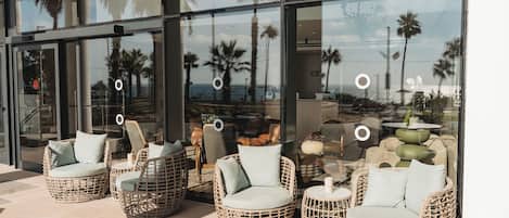 Wicker chairs with cushions outside a building with large windows reflecting palm trees and a view of the ocean.