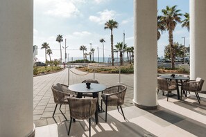 An outdoor patio area with round tables and wicker chairs, palm trees, and a view of the ocean. - ALMERA  (Bat Yam)