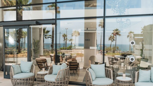 Un área de estar moderna al aire libre con sillas y mesas de mimbre, con vista a una playa y palmeras.