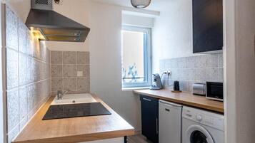 A compact kitchen with a sink, stove, and dishwasher, featuring a window and tiled walls.