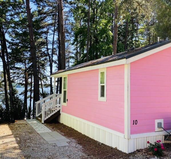 Una cabaña rosa con un porche blanco y una ventana pequeña, rodeada de árboles altos y una masa de agua.