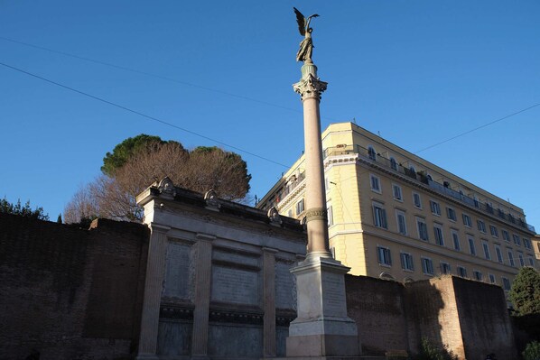 A classical column with a statue on top, a large building with multiple windows, and a clear blue sky. - Bonnavista inn (Rome)