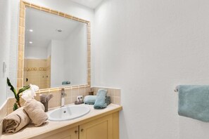 Un baño con un lavabo blanco, tocador color beige y un espejo grande. Hay toallas y una planta en el tocador.