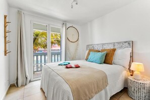 Un dormitorio con cama, lámpara de noche, ventana con cortinas, decoración en la pared y vistas a palmeras fuera.