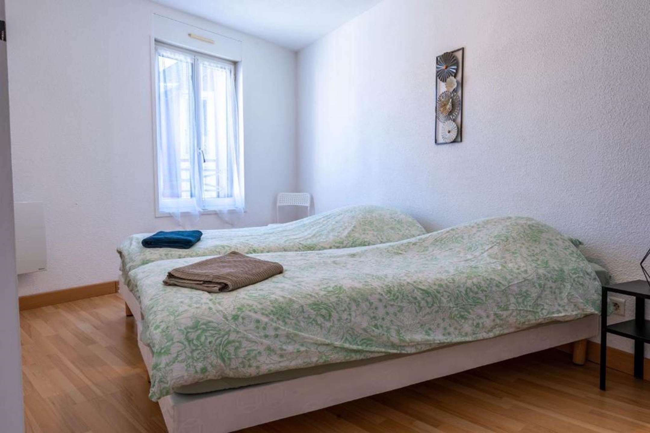 A bedroom with two beds, a window with curtains, a chair, and a wall-mounted decoration.