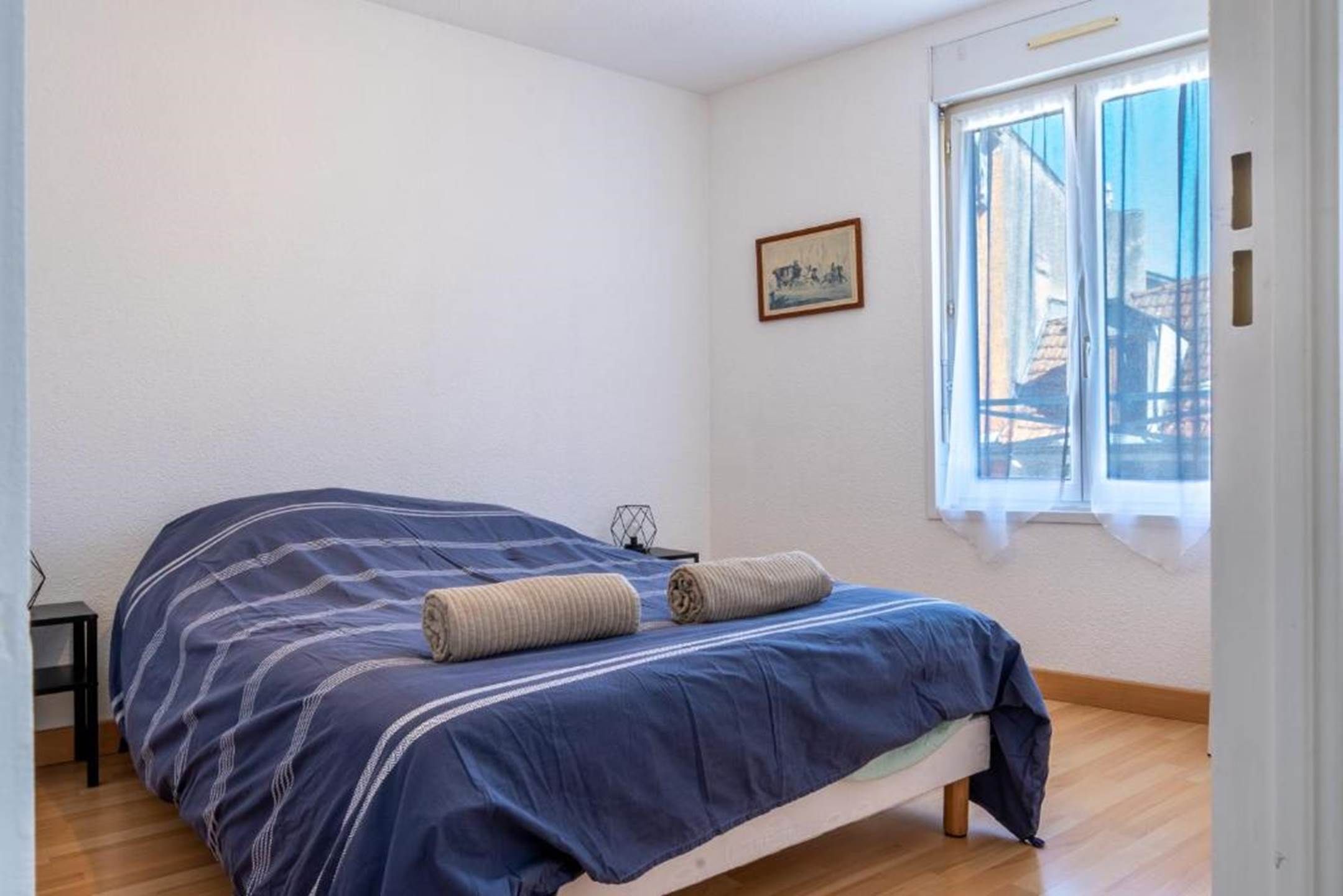 A single-bed room with a wooden floor, a window with a view of buildings, and a small table with a chair.