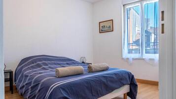 A single-bed room with a wooden floor, a window with a view of buildings, and a small table with a chair.