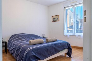 Une chambre avec un lit simple, un plancher en bois, une fenêtre donnant sur des immeubles et une petite table avec une chaise.
