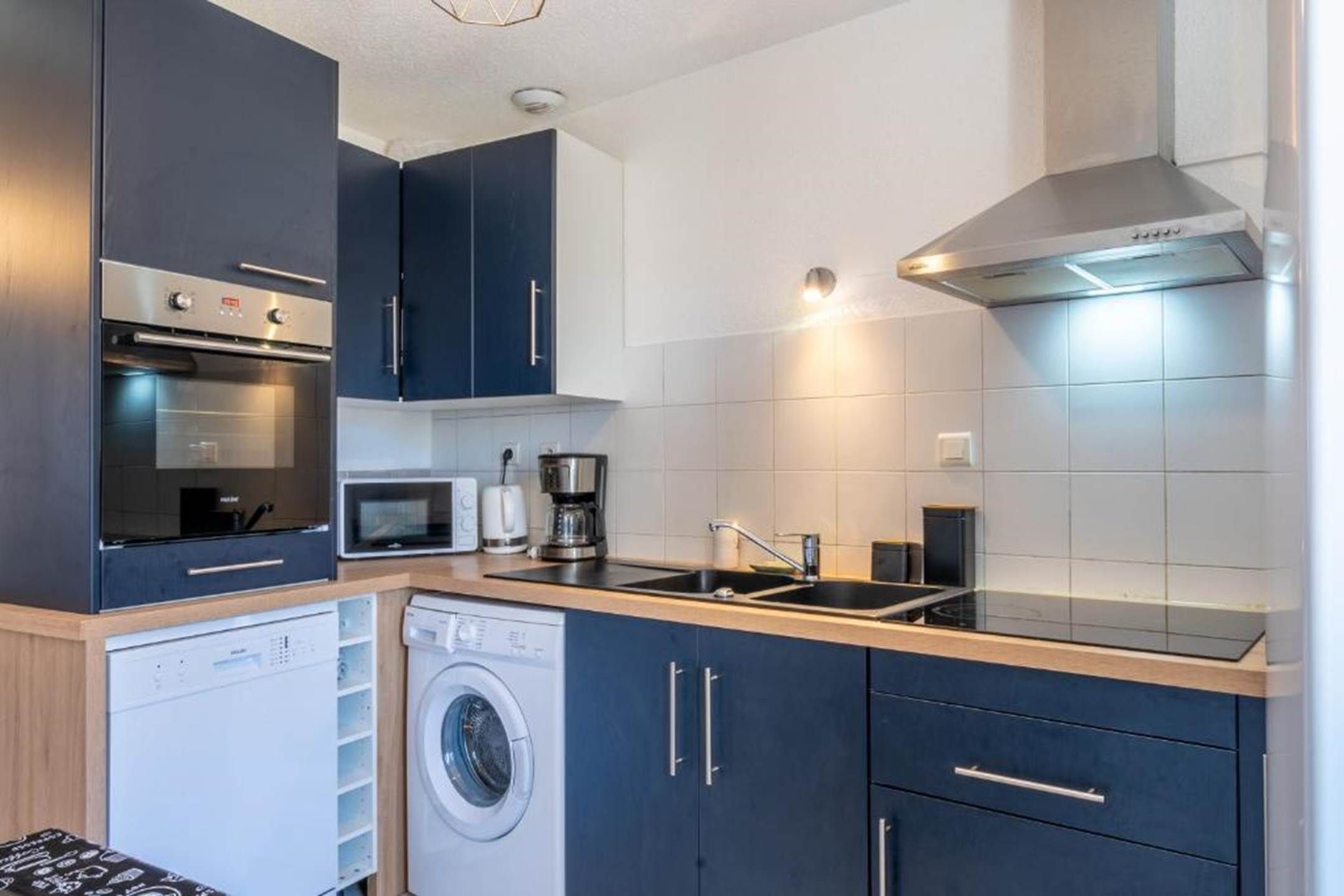 A compact kitchen with a washing machine, microwave, coffee maker, and a stainless steel range hood.