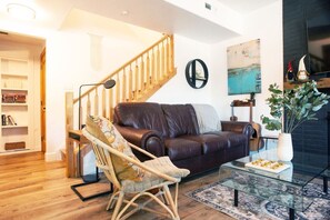 A living room with a leather sofa, a wooden chair, a glass coffee table, a staircase, and a bookshelf.