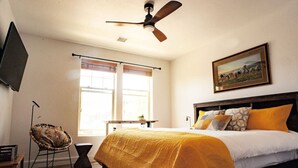 Una camera da letto con un letto, una ventola a soffitto, una finestra con tende, una sedia e un quadro appeso al muro.