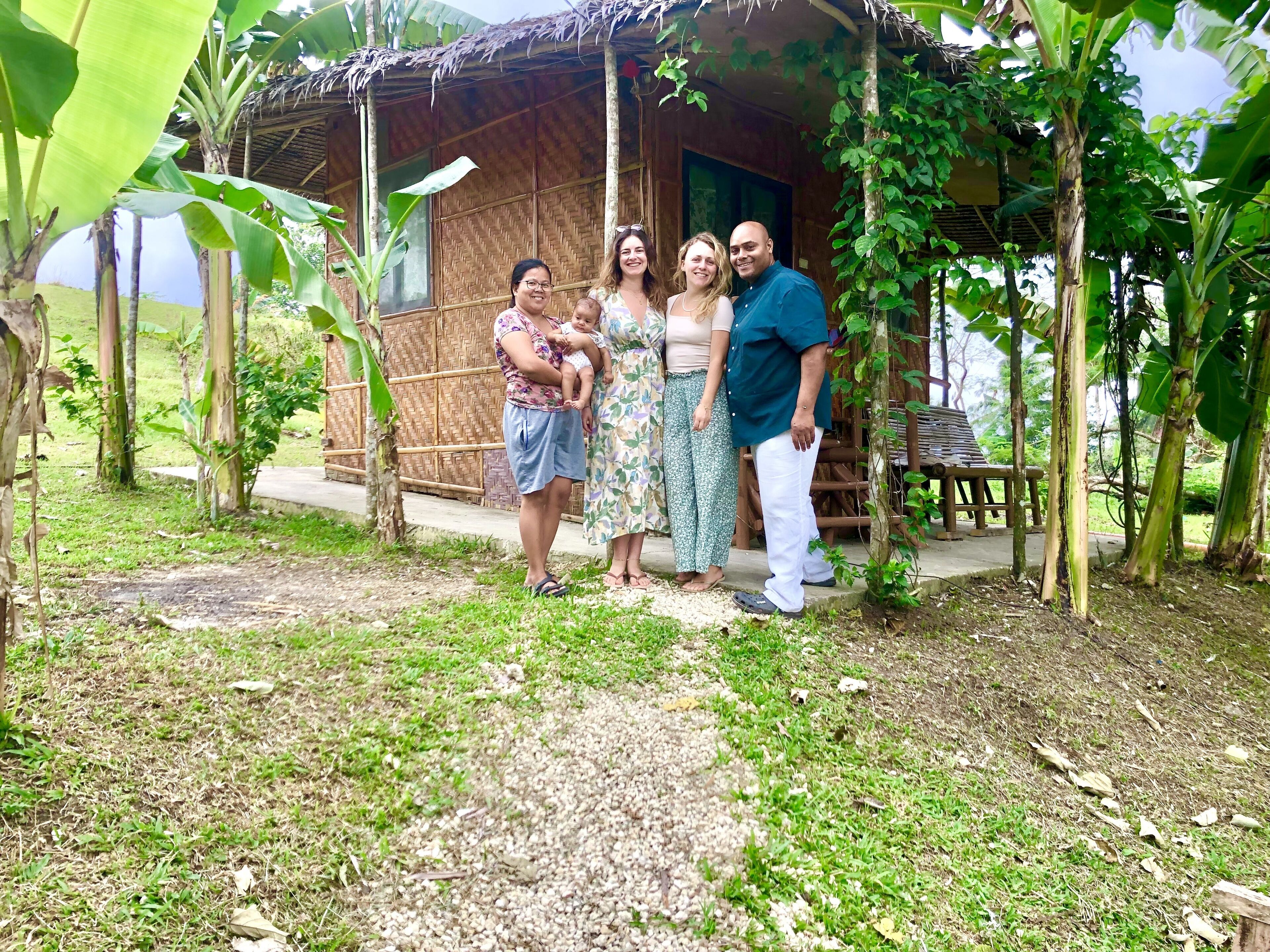 A family standing in front of a rustic wooden house with a thatched roof, surrounded by lush greenery.