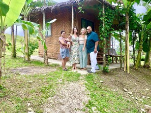 A family standing in front of a rustic wooden house with a thatched roof, surrounded by lush greenery.